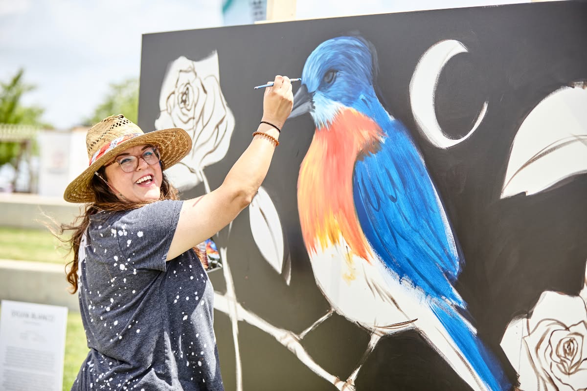 woman painting a blue, yellow and orange bird.